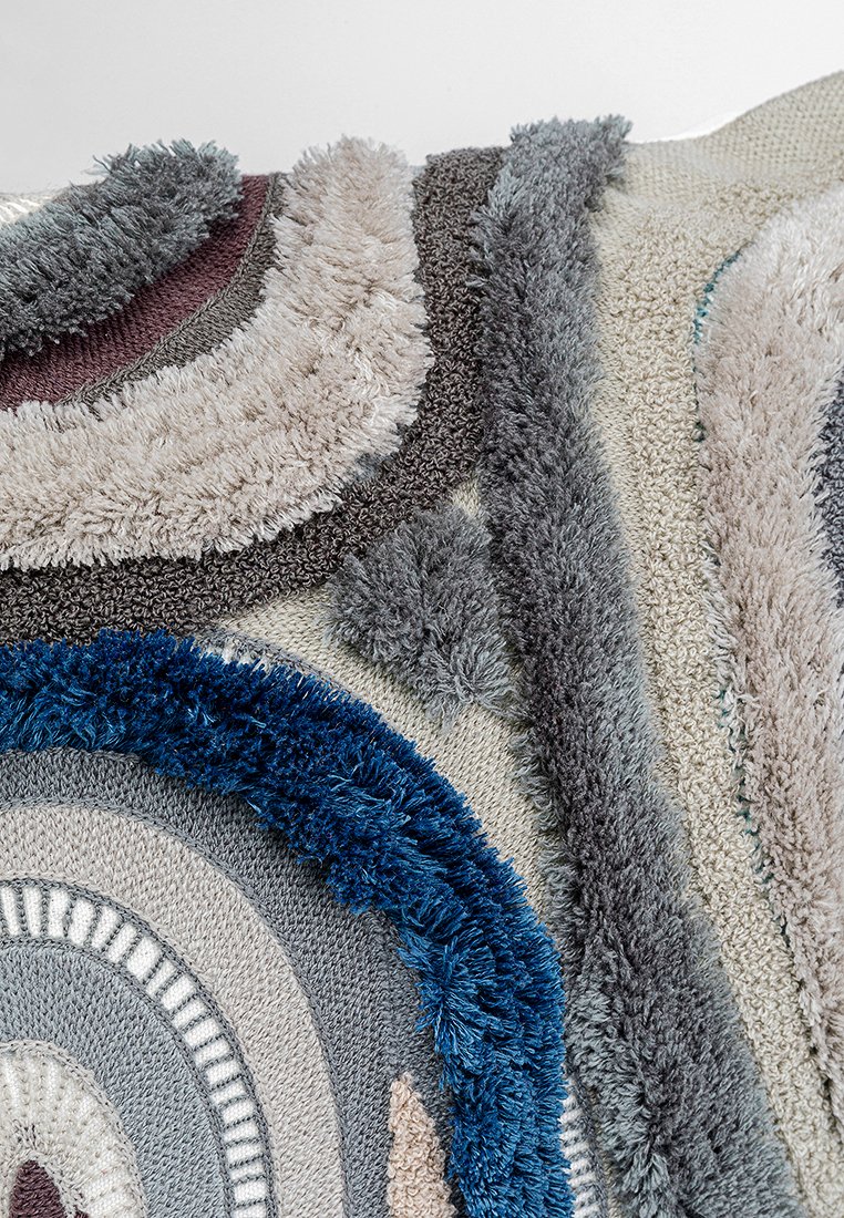 Mehrfarbiger Teppich mit kreisförmigen Mustern in Blau, Grau, Creme und Beige. Dichte, strukturierte Fasern schaffen Tiefe und Kontrast.