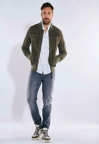 Olivgrüne Wildleder-Bomberjacke mit grauen Akzenten, gestreiftes weißes Hemd, graue Jeans und High-Top-Sneaker mit grauen und cremigen Akzenten.