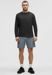 Uomo in piedi contro uno sfondo semplice, indossando una maglietta nera a maniche lunghe, pantaloni grigi, calze bianche e scarpe da ginnastica nere.
