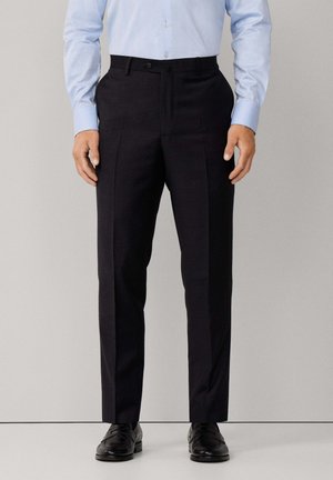 Mann trägt schwarze formelle Hose, hellblaues Hemd mit Knöpfen und schwarze Lederschuhe und steht vor einem einfarbigen Hintergrund.