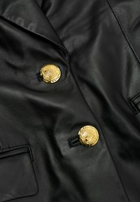 Szczegół czarnej skórzanej kurtki z dwoma błyszczącymi, złotymi, tłoczonymi guzikami oraz przeszywaną klapką kieszeni.