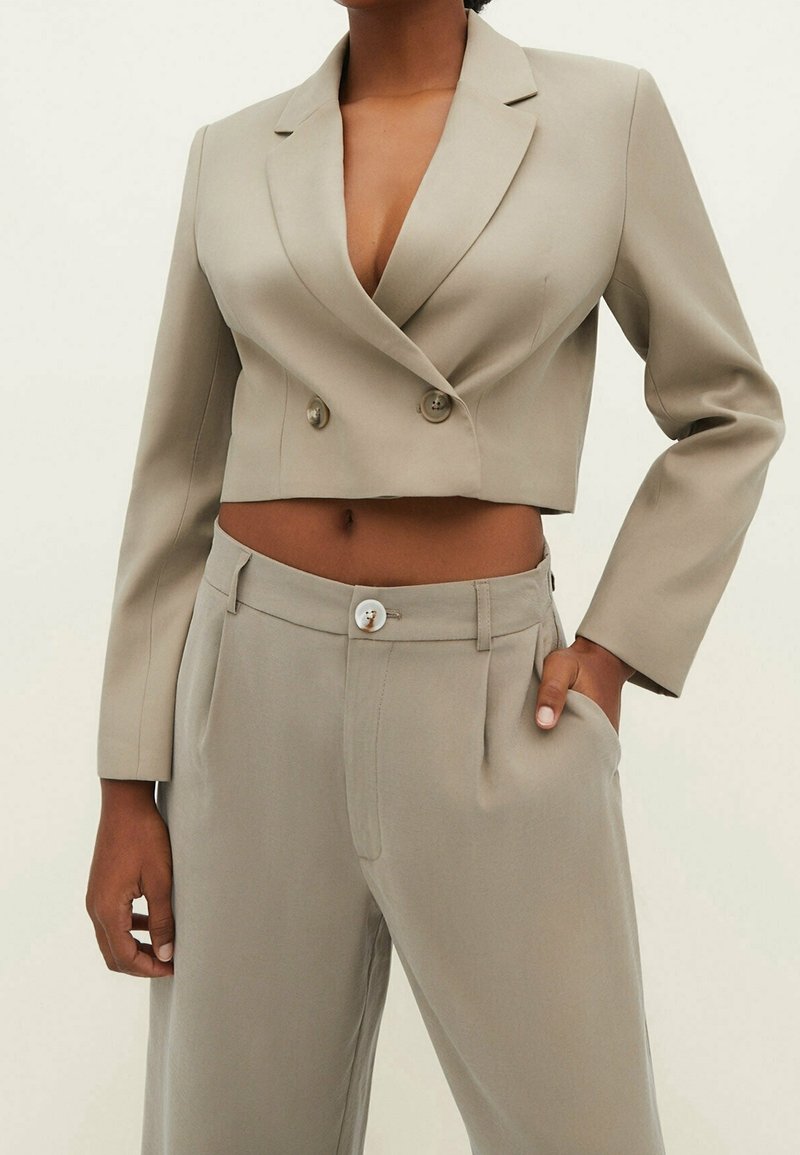 Blazer beige court à double boutonnage avec revers crantés, associé à un pantalon assorti avec une fermeture à boutons sur le devant et des plis.