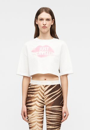 Młoda kobieta ubrana w białą krótka bluzkę z logo ust "Just Cavalli" oraz legginsy z wysokim stanem w paski tygrysa, stojąca na jednolitym tle.