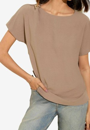 Femme portant un haut taupe à manches courtes ample avec un détail boucle à l'épaule, et un jean bleu clair, main dans la poche.
