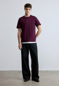 Maroon cotton t-shirt with a crew neck and embroidered logo. Paired with wide-leg dark denim jeans. Smooth texture, casual design.