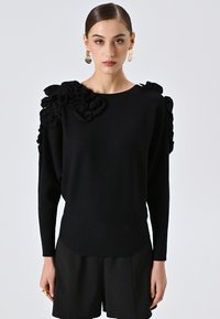 Maglione nero a maglia con dettagli arricciati sulla spalla sinistra, maniche lunghe e scollatura rotonda. Abbinato a pantaloni corti neri.
