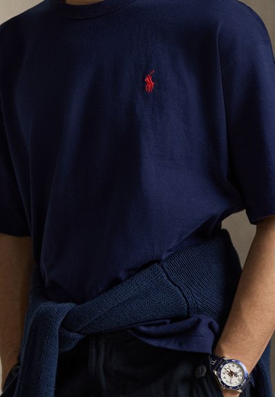 Μπλε σκούρο πόλο πουκάμισο με κόκκινο κεντημένο λογότυπο, ασορτί πλεκτή μπλούζα δεμένη στη μέση και μπλε ρολόι με λευκό καντράν στον αριστερό καρπό.