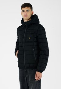 Giubbotto a vento nero con zip frontale, cappuccio e dettagli cuciti; presenta una tasca sul petto e tasche laterali con una texture liscia.