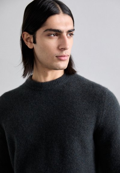 Pull en laine gris foncé avec un col rond, surface texturée et silhouette ajustée, porté par un mannequin aux longs cheveux foncés.