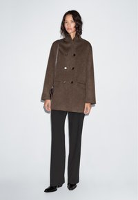 Brown wool peacoat with a high collar, double-breasted buttons, and side pockets. Paired with black trousers and heeled boots.