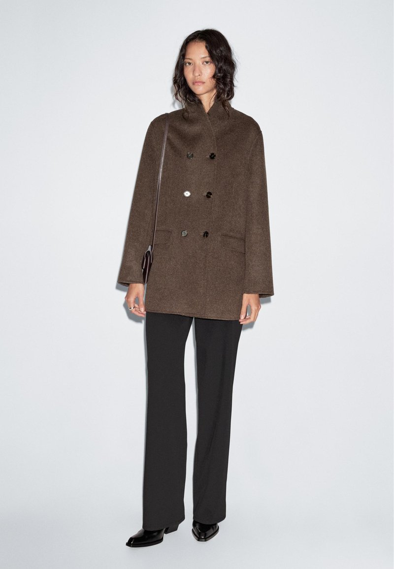 Brown wool peacoat with a high collar, double-breasted buttons, and side pockets. Paired with black trousers and heeled boots.