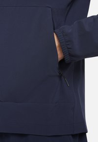 Marineblauwe jas met een ritszak en elastische manchetten, gemaakt van een gladde stof met een matte afwerking en een minimalistisch design.