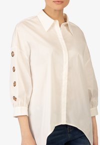 Camicia bianca con colletto e polsini abbottonati, dotata di bottoni in tonalità oro sulle maniche. Vestibilità ampia, orlo curvo e tessuto leggero.