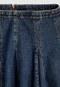 Gros plan d'une jupe en denim bleu foncé avec des coutures verticales, un détail de plis et une fermeture éclair en laiton à la taille.