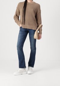 Jasnobrązowy sweter z dzianiny, niebieskie dżinsy o prostych nogawkach i białe sneakersy na platformie. Trzyma małą beżową torebkę na ramię. Prosty design.