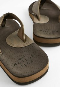Brūni flip-flopi ar strukturētu kāju daļu, mīkstu auduma siksnu un kontūrētu gumijas zoli. Iespējama "NEXT MOTION FLEX" zīmola marķējums melnā krāsā.