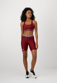 Top corto atlético de color marrón con el logo de Under Armour, combinado con pantalones cortos de tiro alto a juego. Zapatillas negras con detalles blancos. Textura de tela suave.