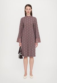 Langärmliges, mauvefarbenes Kleid mit einem Diamantmuster, das über einen hohen Kragen und ausgestellte Ärmelabschlüsse verfügt. Das Model hält eine kleine schwarze Handtasche.