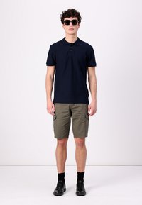 Námořnické polo tričko s krátkým rukávem, khaki cargo kraťasy, černé sluneční brýle, černé ponožky a černé boty, stojící na jednobarevném pozadí.