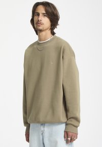 Sweatshirt vert olive avec un col rond, des manches longues et des poignets côtelés ; détail logo doré sur la poitrine ; porté par-dessus un T-shirt blanc.