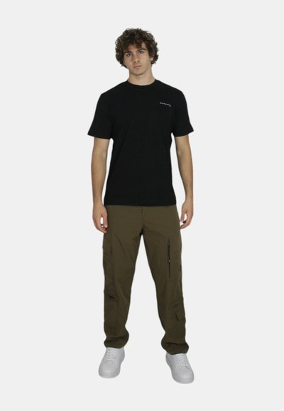 T-shirt nera in cotone con maniche corte, abbinata a pantaloni cargo verde oliva con multiple tasche e cerniere, completata da sneakers bianche.