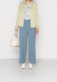 Light green button-up shirt over a patterned blue and white top, paired with wide light blue pants and off-white heeled ankle boots. Holding a pale handbag.