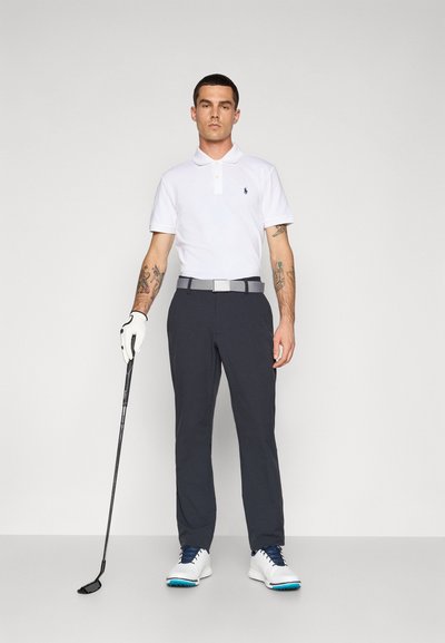 Valkoinen polo-paita, jossa on pieni logo, harmaa vyö, tummanharmaat housut, valkoiset kengät sinisillä yksityiskohdilla ja kädessä golfmaila.
