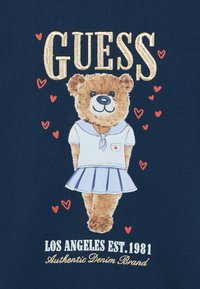 Το γραφικό παρουσιάζει μια καφέ αρκούδα παιχνιδιού με μπλε φούστα και λευκό πουκάμισο με ναυτικό γιακά. Το κείμενο γράφει «GUESS» σε χρυσό χρώμα πάνω και τις λεπτομέρειες της μάρκας κάτω.