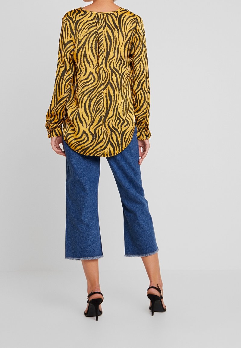 Donna in piedi di spalle, indossa una blusa a stampa animale gialla e nera, jeans blu a vita alta e sandali neri con tacco alto.