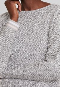 Person, der einen strukturierten grauen und weißen Strickpullover mit gestreiften Bündchen trägt und mit einer Hand das Kinn nahe dem Gesicht stützt.