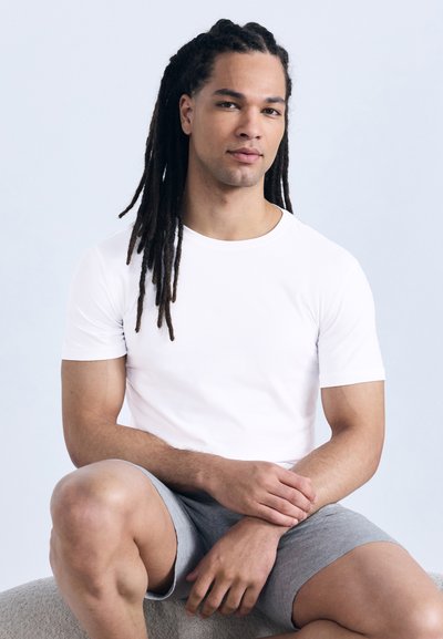 Νέος άνδρας με μακριά ντραντλόκ, φορώντας λευκό μπλουζάκι και γκρι σορτς, καθισμένος με τα χέρια ακουμπισμένα σε ένα γόνατο, μπροστά σε ένα ανοιχτόχρωμο φόντο.