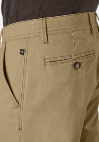 Khaki-Hosen mit strukturierter Oberfläche, ausgestattet mit einer Gesäßtasche, die mit einem Knopf gesichert ist, und einem kleinen schwarzen Etikett in der Nähe des Taillenbunds.