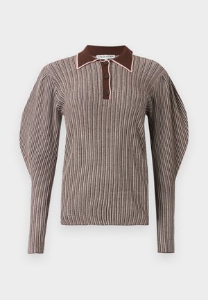 Pikk naine pruunide triipudega kaelusega pika varrukaga toppi, musta teksase seeliku ja mustade teravate kontsakingadega, seisab käed lõdvalt.