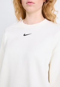 Sudadera de color crema con cuello redondo, que presenta un pequeño logo negro de Nike en el pecho. Textura suave, corte relajado.