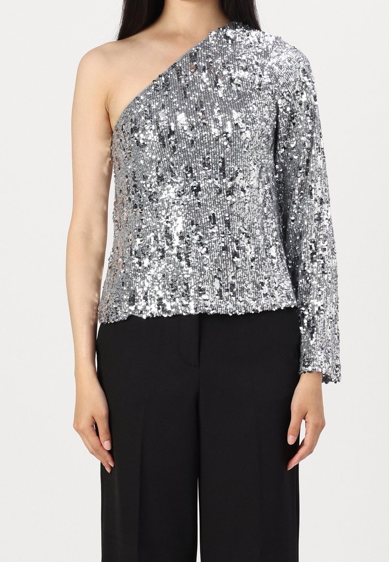 Femme portant un top asymétrique argenté à manches longues avec sequins, associé à un pantalon noir à jambes larges, sur un fond uni.