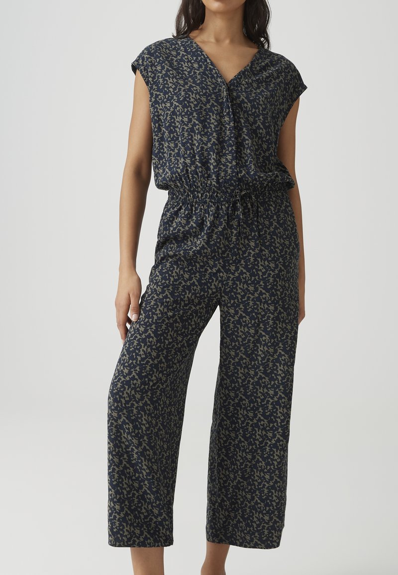 Frau trägt ärmellosen marineblauen Overall mit olivfarbenem abstraktem Muster, elastischem Bund und weitem Bein vor einfarbigem Hintergrund.