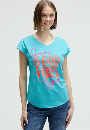 Junge Frau mit kurzen hellbraunen Haaren, die ein türkisfarbenes T-Shirt mit V-Ausschnitt und dem Aufdruck „Good Vibes“ in großen roten und pinken Buchstaben trägt.