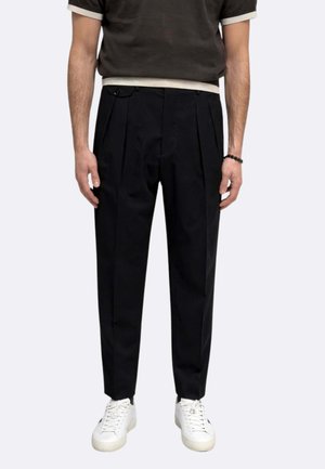 Uomo con pantaloni neri a pieghe, scarpe da ginnastica bianche, camicia nera con maniche a bordo bianco, in piedi contro uno sfondo chiaro e semplice.