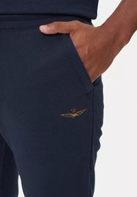 Pantaloni della tuta blu navy con una finitura testurizzata, caratterizzati da un logo ad ali d'oro ricamato sopra la tasca anteriore.