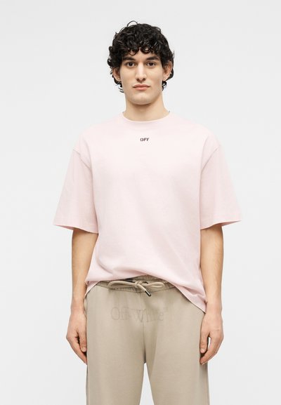 Νεαρός με σγουρά μαλλιά, ντυμένος με ροδακινί φαρδιά μπλούζα με το κείμενο "OFF" και μπεζ παντελόνι με κορδόνι και διακριτική επωνυμία "Off-White".