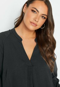 Mörkgrå blus med v-ringning, lätt textur och långa ärmar. Har subtila plisseringar och sida fickor.