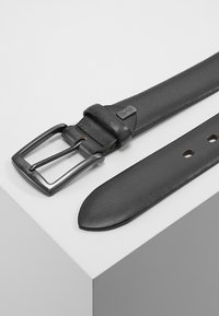 Cinturón de cuero negro con una hebilla metálica rectangular de color plata, textura suave, extremo afilado y varios agujeros de ajuste a lo largo de la correa.
