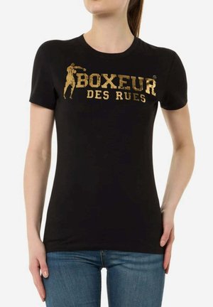 Frau trägt ein schwarzes, eng anliegendes T-Shirt mit goldfarbener Aufschrift "Boxeur des Rues" und Boxersilhouette, kombiniert mit blauen Jeans.