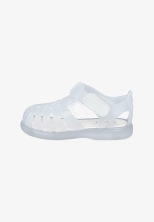 Sandalias blancas translúcidas con punta cerrada y lados abiertos, que cuentan con una correa de Velcro y un diseño ranurado para ventilación.