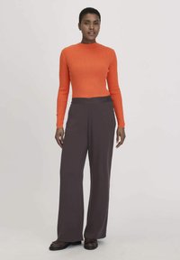 Orange gerippter Strickpullover mit langen Ärmeln, kombiniert mit weit geschnittenen, dunkelbraunen Hosen. Das Model steht mit den Händen in den Hüften und trägt braune Schuhe.