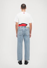 Camiseta blanca con un detalle gráfico en rojo y negro en la parte posterior, combinada con jeans de pierna ancha de color azul claro y zapatillas negras.
