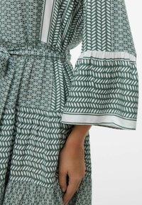 Vestido verde con estampado geométrico variado, que presenta una cintura ceñida, mangas acampanadas y una falda en capas con detalles blancos.