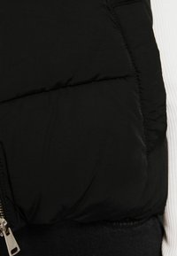 Black puffer vest with a smooth, matte texture. Features a silver zipper and a subtle seam design, layered over a white ribbed top.