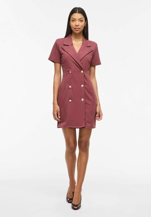 Kvinne står iført kortermet, støvrosa blazerkjole med seks gullknapper, kombinert med svarte peep-toe hæler mot hvit bakgrunn.