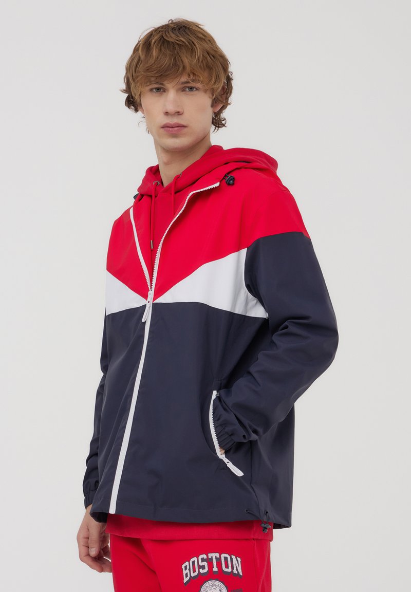 Giacca a blocchi di colore con pannelli rossi, bianchi e blu navy, realizzata in un materiale impermeabile e liscio. Cappuccio con coulisse e chiusura con zip.